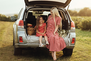 A mother and her young daughter, both wearing matching red and white dresses, sit smiling in the open trunk of a silver SUV parked in a golden field