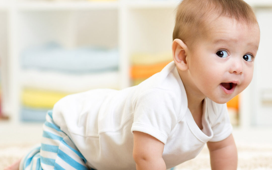 A wide-eyed baby wearing a white shirt and blue striped pants crawls on a soft rug with a surprised, happy expression
