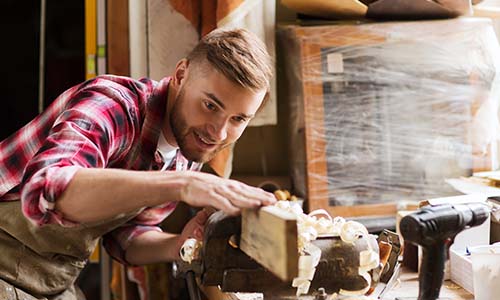 A man carefully shapes a wooden piece using tools in his workshop, surrounded by various woodworking equipment
