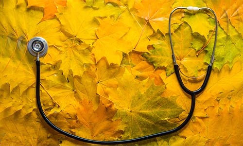 A stethoscope rests on a colorful pile of autumn leaves, blending medical and seasonal themes